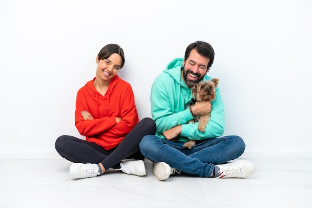 Young caucasian couple sitting on the floor with their pet isolated on white background keeping the arms crossed while smiling