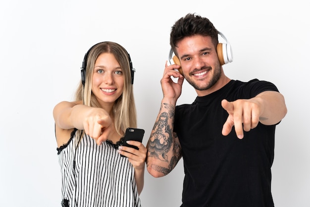 Young caucasian couple on white listening music and pointing to the front