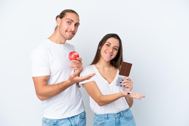 Young caucasian couple with chocolat and apple isolated on white background pointing back and presenting a product