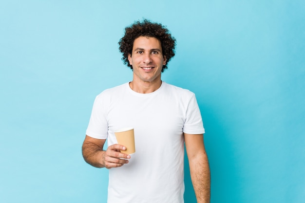 Young caucasian curly man holding a takeaway coffee happy, smiling and cheerful.