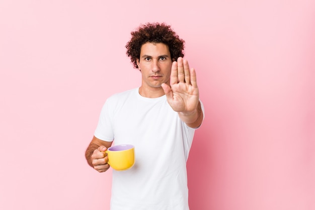 Young caucasian curly man holding a tea cup standing with outstretched hand showing stop sign, preventing you.