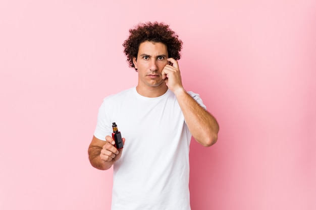 Young caucasian curly man holding a vape pointing his temple with finger, thinking, focused on a task.