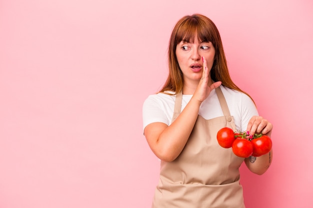 Young caucasian curvy woman cooking at home holding tomatoes isolated on pink background is saying a secret hot braking news and looking aside