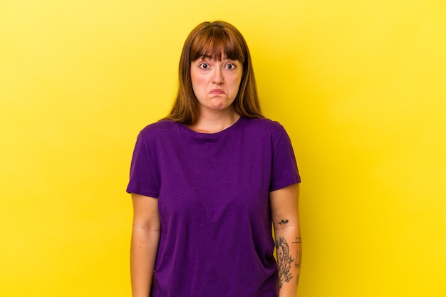 Young caucasian curvy woman isolated on yellow background shrugs shoulders and open eyes confused.