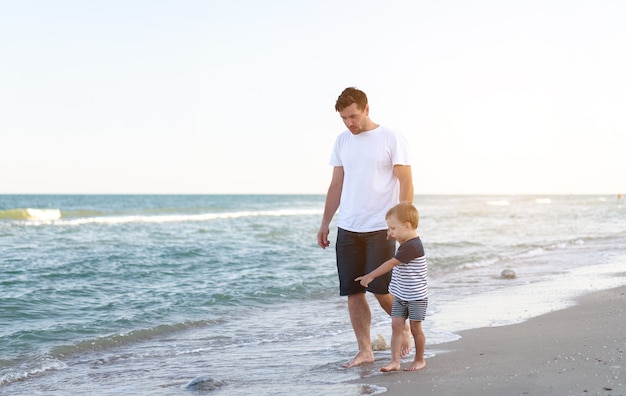 Young Caucasian dad with little son walk warm summer day along the sea coast