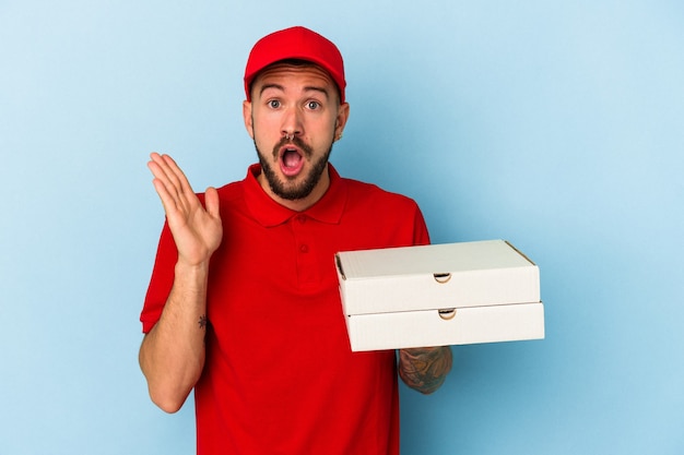 Young caucasian delivery man with tattoos holding pizzas isolated on blue background  surprised and shocked.