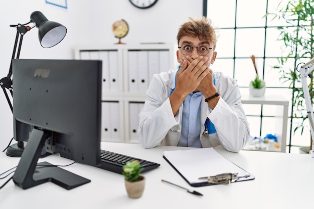 Young caucasian doctor man working at the clinic shocked covering mouth with hands for mistake. secret concept.