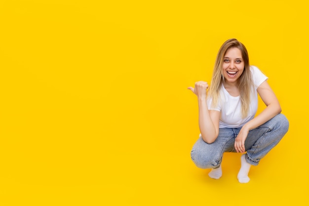 A young caucasian excited smiling blonde woman sitting on her haunches pointing copy empty blank space for text or design with her finger presenting product isolated on bright color yellow wall