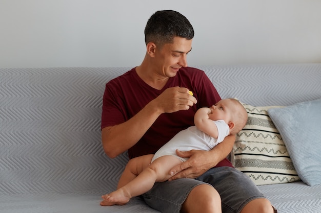 Young Caucasian father gives to baby soother while sitting on sofa in room, looking at her daughter or son with great love, guy wearing maroon t shirt, dad playing with his kid.