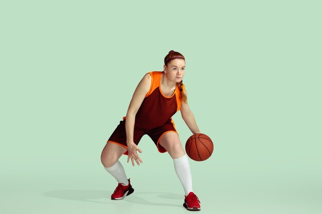 Young caucasian female basketball player in action, motion during gameplay isolated on mint colored background. Concept of sport, movement, energy and dynamic, healthy lifestyle. Training, practicing.