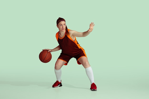 Young caucasian female basketball player in action, motion during gameplay isolated on mint colored background. Concept of sport, movement, energy and dynamic, healthy lifestyle. Training, practicing.