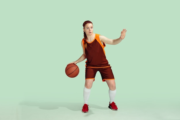 Young caucasian female basketball player in action, motion during gameplay isolated on mint colored background. Concept of sport, movement, energy and dynamic, healthy lifestyle. Training, practicing.