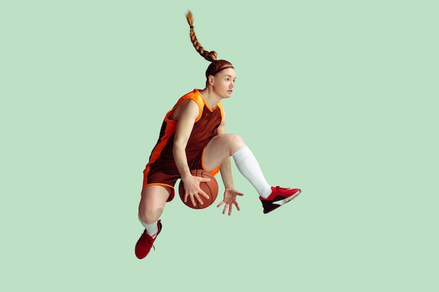 Young caucasian female basketball player in action, motion in high jump isolated on mint colored background. Concept of sport, movement, energy and dynamic, healthy lifestyle. Training, practicing.