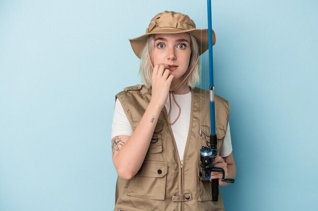 Young caucasian fisherwoman holding a rod isolated on blue background biting fingernails nervous and very anxious