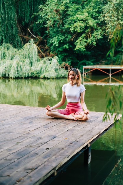 Young caucasian fitness woman doing yoga, meditate outdoors, on wooden pier lake on summer day.