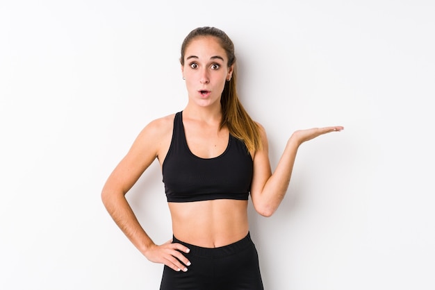 Young caucasian fitness woman posing in a white wall impressed holding copy space on palm.