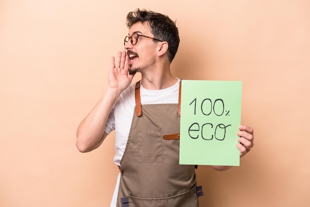 Young caucasian gardener man holding 100 eco placard isolated on beige background shouting and holding palm near opened mouth