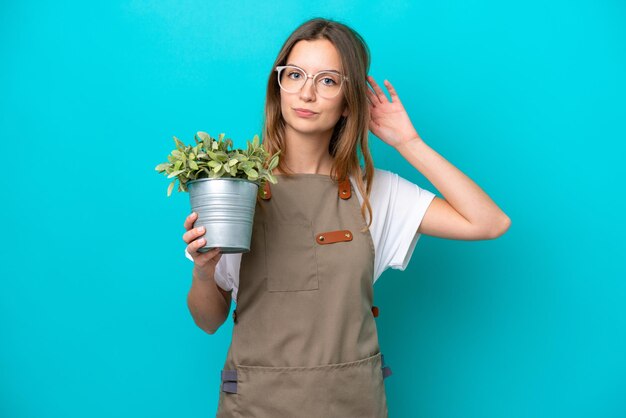 Young caucasian gardener woman holding a plant isolated on blue background having doubts