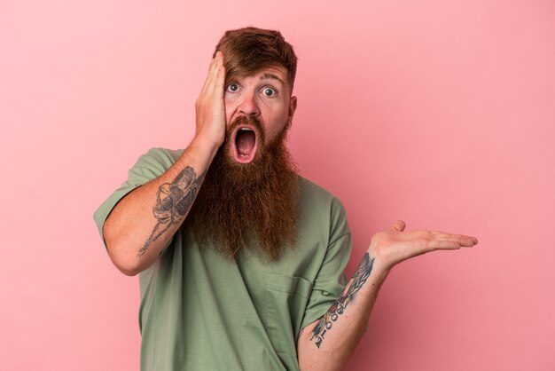 Young caucasian ginger man with long beard isolated on pink background impressed holding copy space on palm.