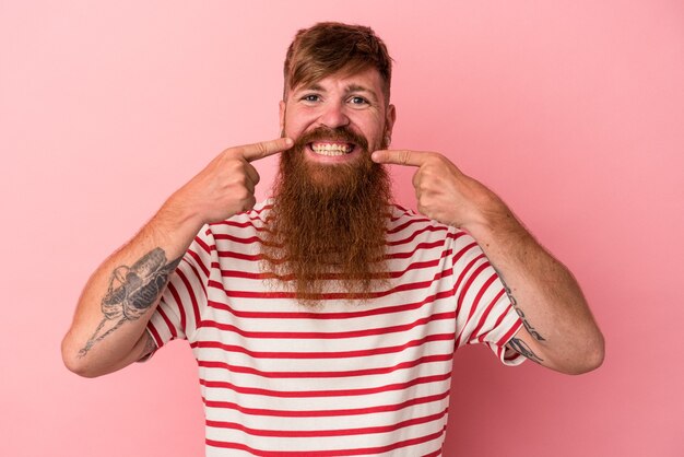 Young caucasian ginger man with long beard isolated on pink background smiles, pointing fingers at mouth.