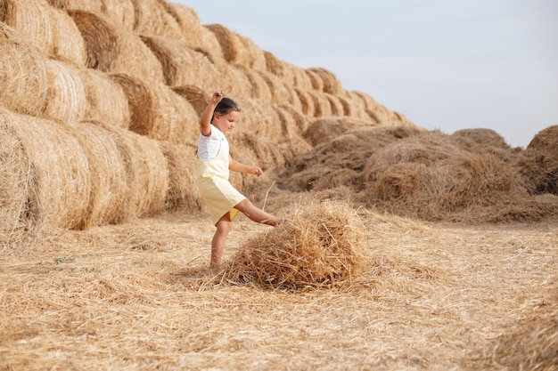 Young Caucasian girl playing and kicking hay with huge heap of hay and big amount of rolled haystacks in background Having fun away from city on field full of golden hay