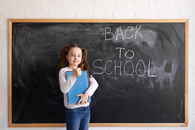 A young Caucasian girl schoolgirl stands against the background of a chalk board The words are written back to school holds several large notebooks in his hands and a thumb up