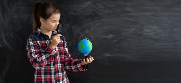 Young caucasian girl schoolgirl or student stands near the chalk blackboard He is holding a globe with the help of a magnifying glass looking for something on the world map Baner