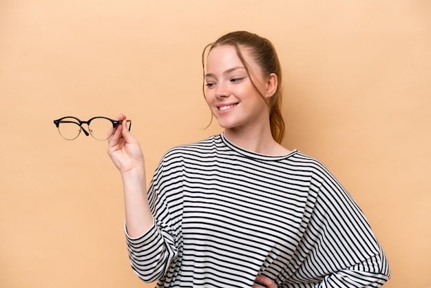 Photo young caucasian girl with glasses isolated on beige background with happy expression