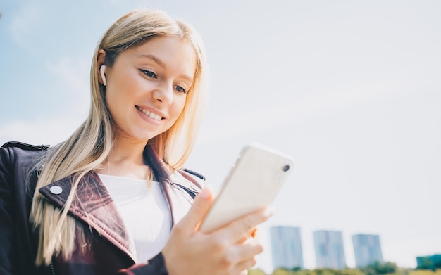 Young caucasian girl with wireless headphones in the park using tablet phone and smiling