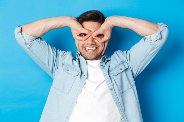 Young caucasian guy showing funny expression, making superhero mask with fingers on eyes, smiling happy, standing over blue background