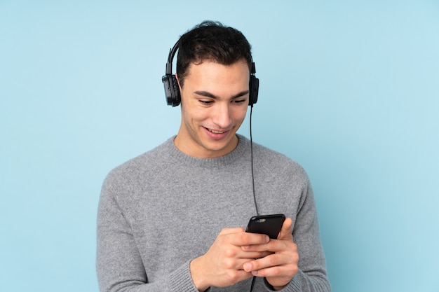 Young caucasian handsome man on blue wall listening music and looking to mobile