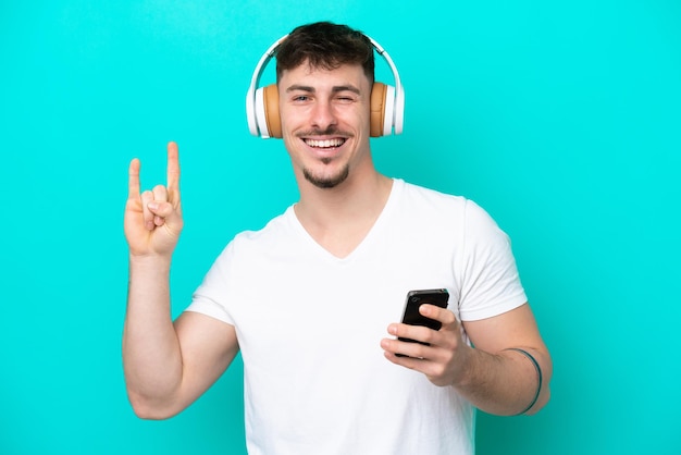 Young caucasian handsome man isolated on blue background listening music with a mobile making rock gesture