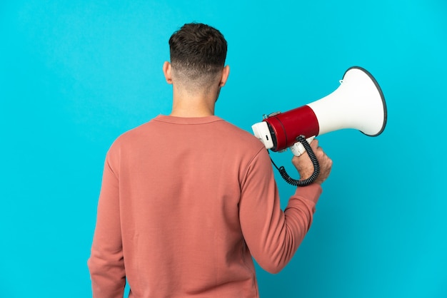 Young caucasian handsome man isolated on blue holding a megaphone and in back position