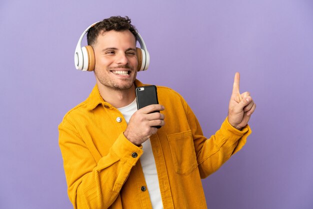 Young caucasian handsome man isolated on purple listening music with a mobile and singing