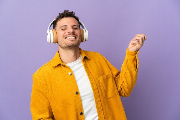 Young caucasian handsome man isolated on purple wall listening music and doing guitar gesture