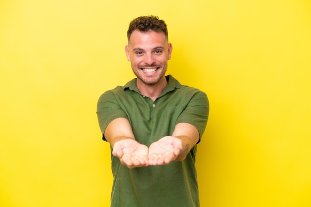 Young caucasian handsome man isolated on yellow background holding copyspace imaginary on the palm to insert an ad