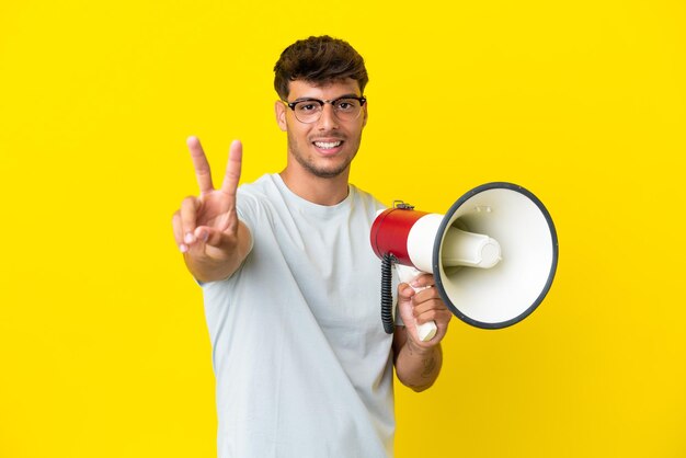 Young caucasian handsome man isolated on yellow background holding a megaphone and smiling and showing victory sign