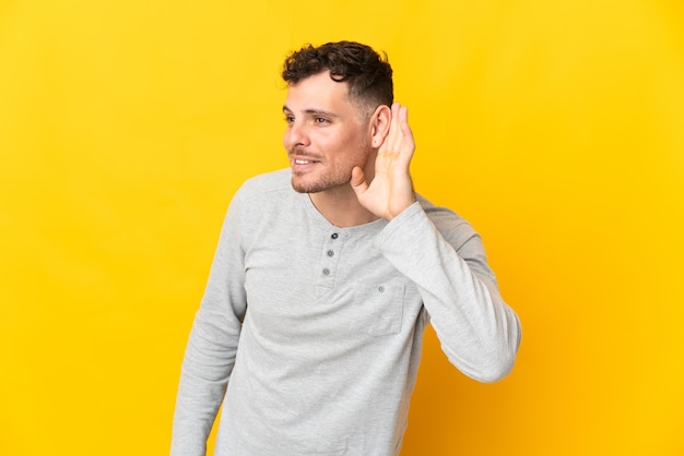 Young caucasian handsome man isolated on yellow listening to something by putting hand on the ear