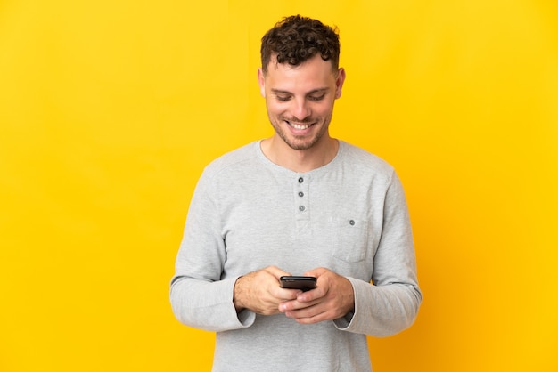 Young caucasian handsome man isolated on yellow wall sending a message with the mobile