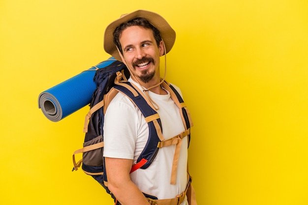 Young caucasian hiker man isolated on yellow background looks aside smiling, cheerful and pleasant.