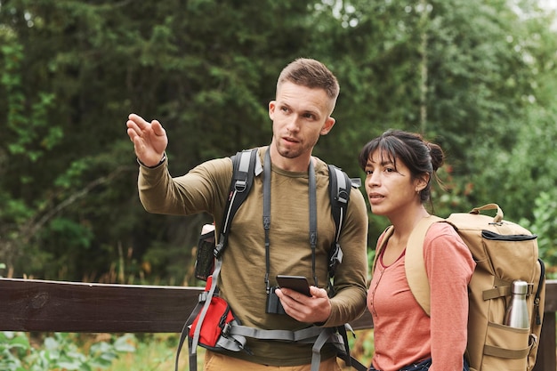 Young caucasian hiker with camera hanging on neck gesturing hand and using phone while explaining ro