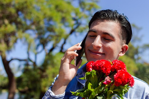 Young caucasian hispanic latin man in love talking to his lover on the phone with his eyes closed holding a bouquet of red flowers near his face outdoors