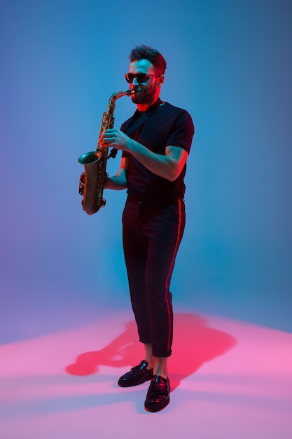 Young caucasian jazz musician playing the saxophone on gradient blue-purple studio background in neon light. Concept of music, hobby, festival. Joyful attractive guy. Colorful portrait of artist.