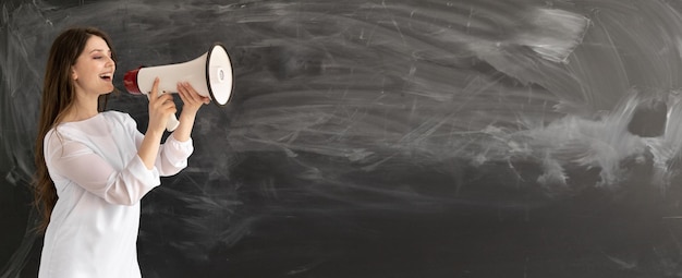 Young Caucasian lady stands against the background of an empty chalk board Holds a loudspeaker and yells loudly into it Announcement concept with a shout Baner