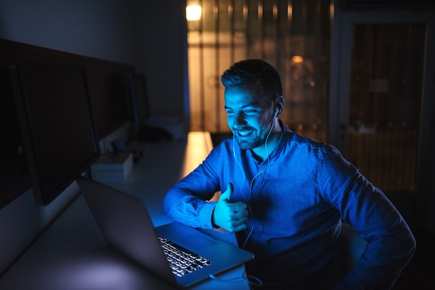 Young Caucasian male employee having video call with boss and showing thumb up while sitting in the office late at night.