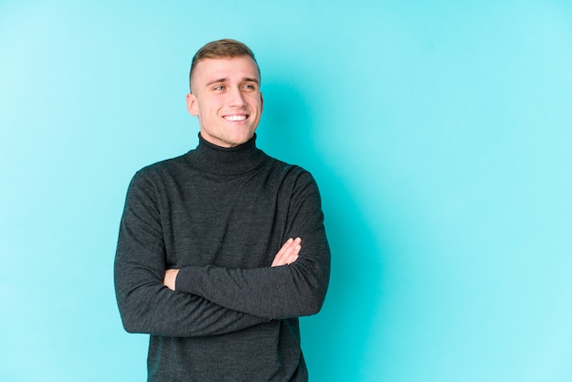 Young caucasian man on a blue wall smiling confident with crossed arms.