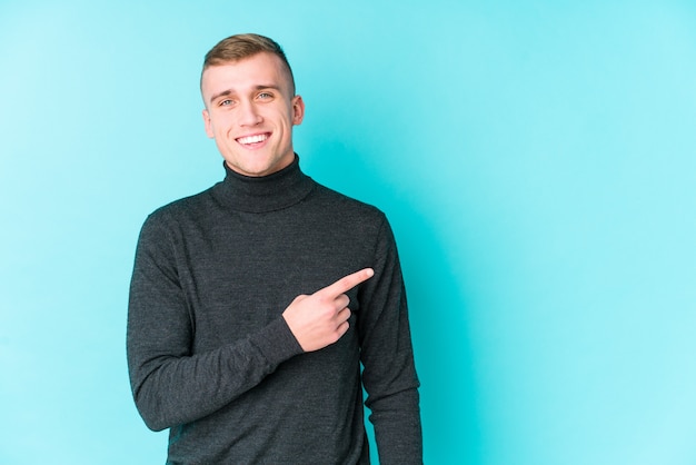 Young caucasian man on a blue wall smiling and pointing aside, showing something at blank space.