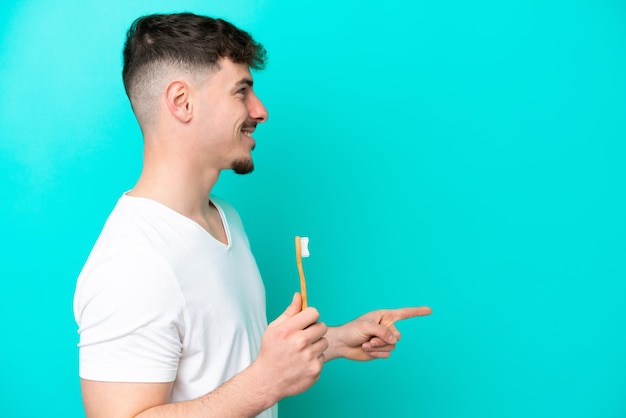 Young caucasian man brushing teeth isolated on blue background pointing to the side to present a product