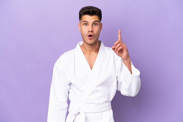 Young caucasian man doing karate isolated on purple background thinking an idea pointing the finger up