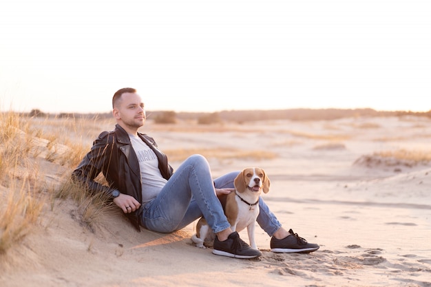 Young Caucasian man dressed black leather jacket and blue jeans sits on sandy beach next to his friend the dog Beagle breed.